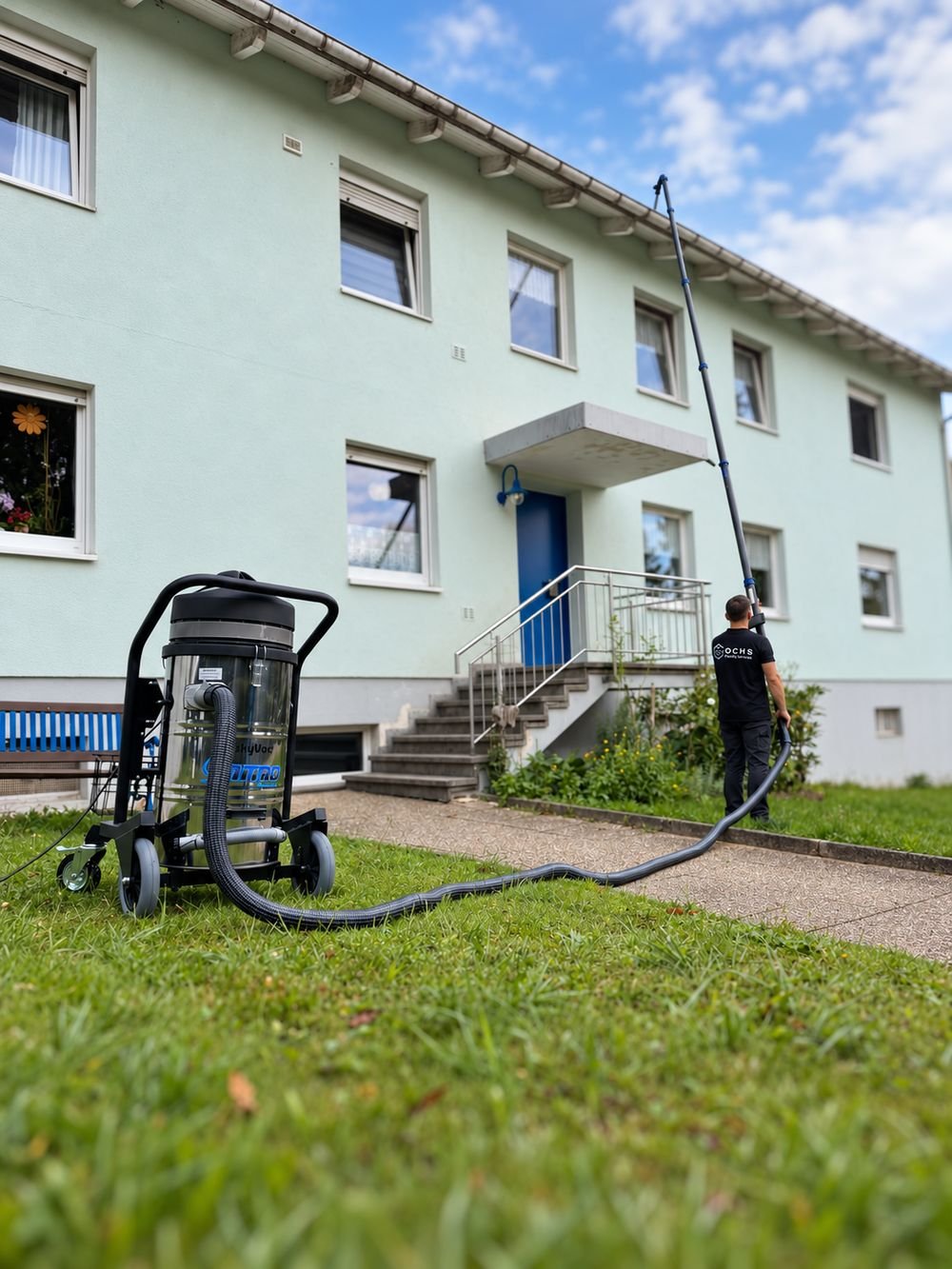 Person im OCHS-Shirt reinigt mit einem Teleskoprohr die Dachrinne eines Mehrfamilienhauses, davor das Sauggerät auf dem Rasen.